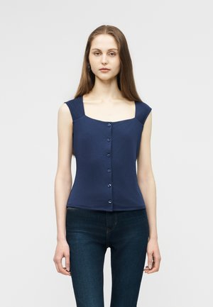 Jeune femme aux longs cheveux raides portant un haut bleu marine sans manches à boutons et un jean bleu foncé, debout contre un fond uni.