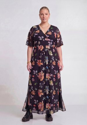 Kvinde iført en sort blomstermaksikjole med gennemsigtige korte ærmer og sorte combatstøvler mod en ensfarvet lys baggrund.