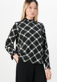 Giovane donna che indossa una blusa a maniche lunghe bianca e nera con collo a volant alto e pantaloni neri, in posa con una mano in tasca.