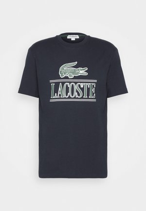 Homme portant un t-shirt Lacoste bleu marine, un jean bleu clair déchiré et des baskets blanches, debout devant un mur clair uni.