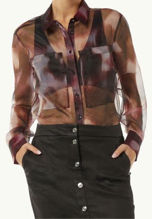 Femme portant un chemisier transparent multicolore à boutons avec des motifs abstraits foncés et une jupe noire taille haute avec des boutons à l'avant et les mains dans les poches.