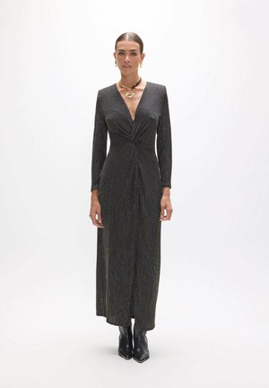 Femme portant une robe noire à manches longues avec des rayures métalliques, un col en V profond et des bottines, debout devant un fond blanc uni.