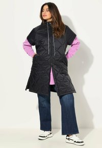 Černá prošívaná bezrukávová bunda s vysokým límcem a šňůrkami, navrstvená přes růžovou košili s dlouhým rukávem a kombinovaná s džíny širokého střihu.