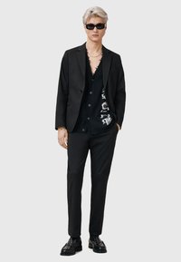Svart kostym med en skräddarsydd kavaj och slim fit byxor, lager på lager över en svart knapp-up cardigan. Glansiga svarta skor och solglasögon fulländar looken.