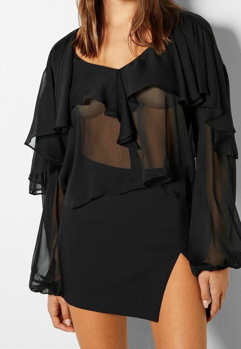 Femme portant un chemisier noir transparent avec des détails de volants et une mini-jupe noire ajustée avec une fente sur la cuisse, visible du cou jusqu'à la moitié de la cuisse.