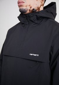Primer plano de una persona que lleva una chaqueta con capucha negra de Carhartt, con cremallera y cordón en el cuello.