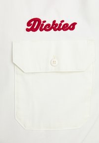 White pocketed shirt featuring red embroidered "Dickies" logo, button closure, and textured fabric with a clean, structured design.