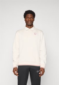 Mand iført en cremefarvet langærmet polo med rødt løvemotiv og sorte Nike-bukser, der står foran en ensfarvet hvid baggrund.