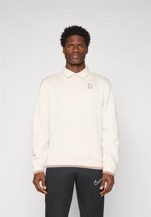 Mand iført en cremefarvet langærmet polo med rødt løvemotiv og sorte Nike-bukser, der står foran en ensfarvet hvid baggrund.