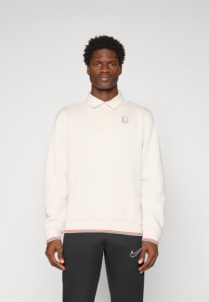 Man in een crèmekleurige lange mouwen polo met een rood leeuw-embleem en zwarte Nike-broeken, staande tegen een effen witte achtergrond.