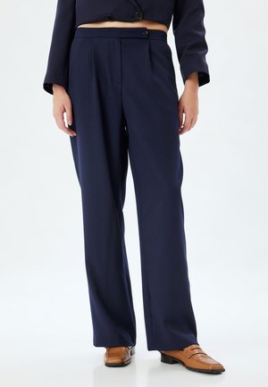Navy brede broek gemaakt van soepele stof, met een schuifsluiting aan de voorkant, plooien en een op maat gemaakte pasvorm. Brown schoenen maken de look af.