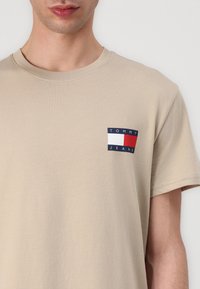 Béžové tričko s krátkym rukávom vyrobené z mäkkej tkaniny, s malou nášivkou v námorníckej modrej a červenej farbe s textom "TOMMY JEANS" naľavo na hrudi.
