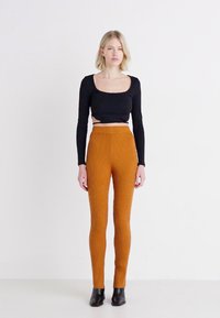 Zwarte cropped langemouwen top met een vierkante halslijn, gedragen met ribbenstof, slim-fit oranje broeken. Model draagt zwarte enkellaarzen. Neutrale achtergrond.