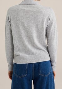 Vaaleanharmaa pitkähihaisen neuleen, jossa on röyhelöhelma ja -hihansuut, yllään korkeavyötäröiset siniset farkut, nähtynä takaa.