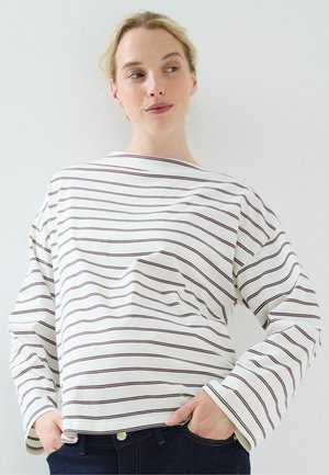 Mujer con cabello rubio que lleva una camisa blanca de manga larga con rayas negras horizontales y vaqueros oscuros, posando con las manos en los bolsillos.
