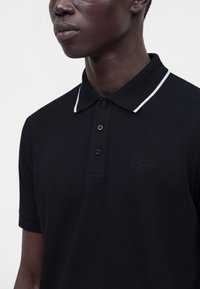 Personne à la peau foncée portant un polo noir avec un liseré blanc au col et un logo brodé sur la poitrine.