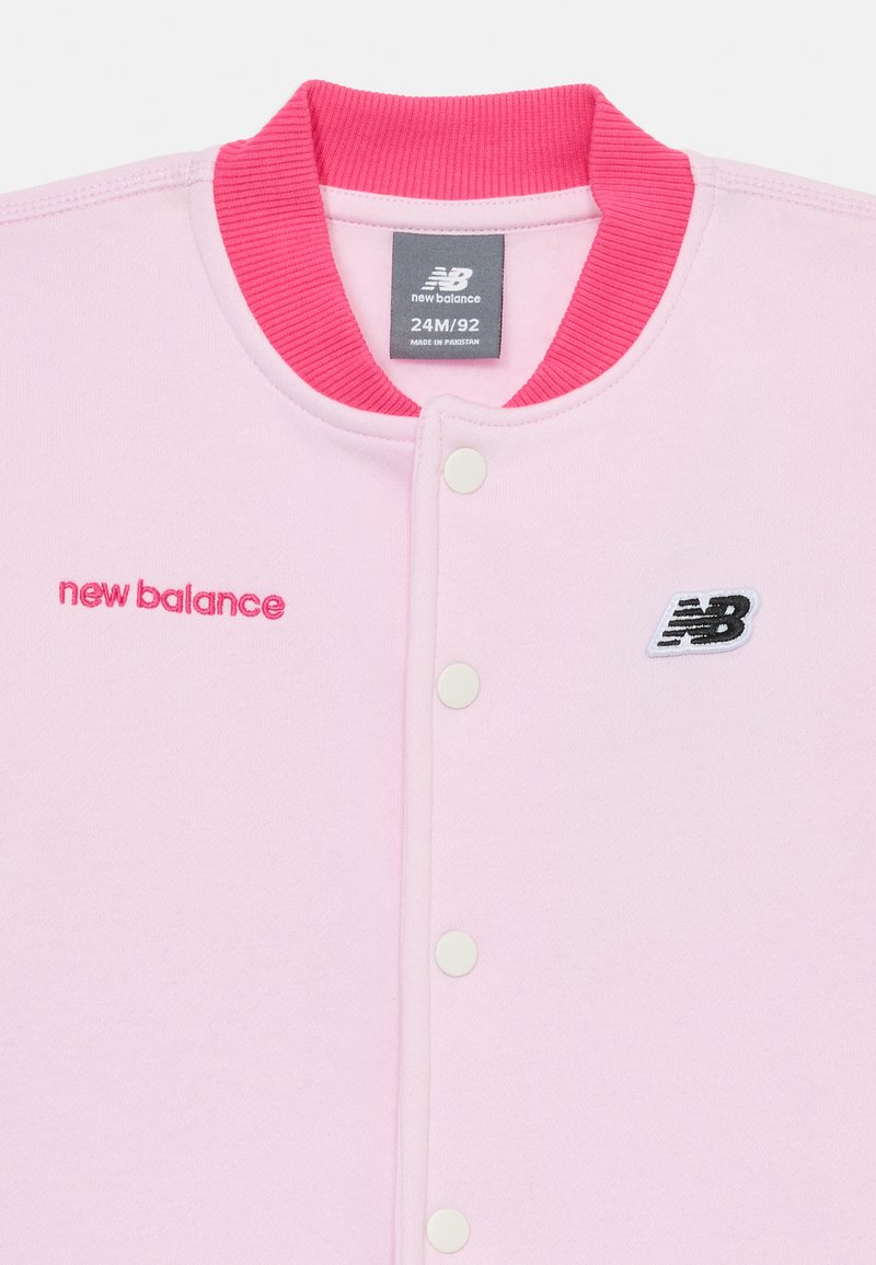 Le sweat-shirt rose de New Balance présente un col rose côtelé, des boutons-pression blancs et un logo noir brodé sur la poitrine.