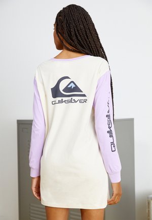 Donna con lunghe trecce che indossa un vestito a maniche lunghe bianco e lavanda con un grande logo Quiksilver sul retro e sulla manica.