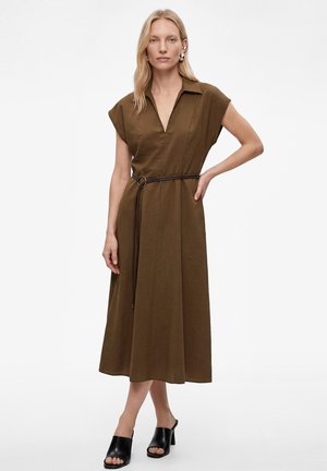 Blonde vrouw in een bruin midi-jurkje met korte mouwen, kraag en dunne riem, zwarte mules met hak en zilveren oorbellen, staand met hand in de zij.