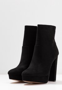 Czarne zamszowe botki za kostkę na wysokim słupkowym obcasie, z okrągłym noskiem i platformą. Gładka tekstura i minimalistyczny design.