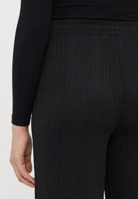 Zwarte broek van textuurstof met verticale strepen, voorzien van een elastische tailleband. Vage glinsterende details versterken het algehele ontwerp.