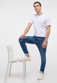 Camiseta blanca de manga corta, jeans ajustados azules y zapatillas blancas. Sentado en una silla blanca con una pierna levantada. Textura suave, estilo casual.