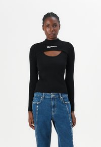 Top lungo a maniche nere con righe, caratterizzato da un colletto alto e un'apertura a forma di chiave, abbinato a jeans blu arricchiti da borchie lungo le cuciture.