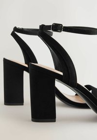 Sandales à talons hauts noires avec un talon bloc, présentant une combinaison de textures lisses et en daim, une bride de cheville ajustable et un design à bout ouvert.