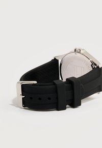 Πίσω όψη ενός ρολογιού χειρός με μαύρο λουράκι σιλικόνης και ασημένια αγκράφα σε λευκό φόντο.