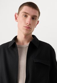 Black zip-up jacket with a textured fabric, sharp collar, and front pocket. Worn over a light gray sweater, showcasing a modern design.