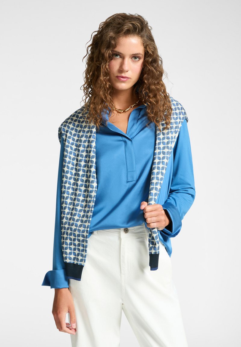 Polo shirt blu a maniche lunghe con colletto. Un maglione con motivo cade sulle spalle. Pantaloni bianchi su misura completano l'outfit.