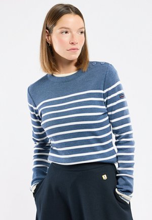 Femme portant un haut à manches longues rayé bleu et blanc et une jupe sombre, les mains dans les poches, regardant légèrement sur le côté.
