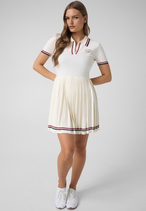 Mujer de pie con las manos detrás de la espalda, lleva un vestido blanco plisado con cuello, puños de las mangas y dobladillo a rayas, combinado con zapatillas blancas.