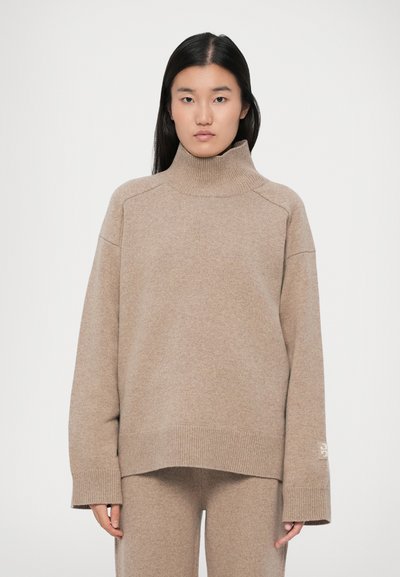 Donna con lunghi capelli neri che indossa un maglione a collo alto beige a coste e pantaloni coordinati su uno sfondo neutro.