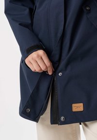 Roka roke zapira zadrgo mornarsko modre jakne z zapenjalnimi gumbi in rjavim usnjenim usnom Swedemount logotipom.