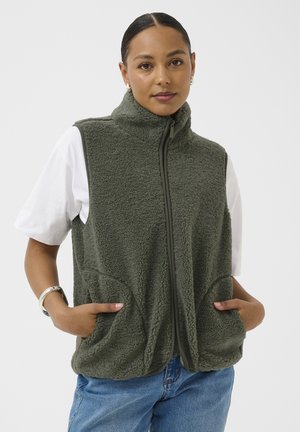 Kobieta ubrana w oliwkowozieloną polarową kamizelkę na zamek, na białej koszulce z krótkim rękawem, z rękami w kieszeniach kamizelki, w połączeniu z niebieskimi dżinsami.