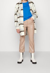 Camisa de cuadros ligera y amplia en crema, azul y negro, combinada con un top azul de cuello redondo y pantalones de cuero sintético beige. Botines blancos de tobillo.