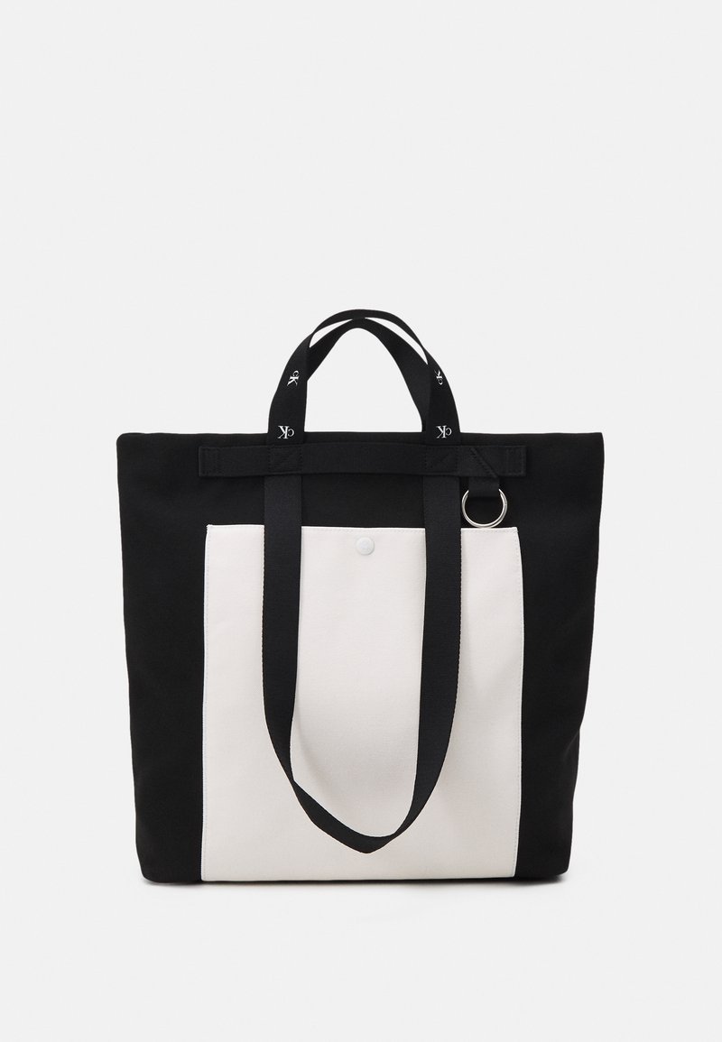 Canvas tote bag in black with a white front panel. Features long black straps with logo accents and a silver metal ring on the side.