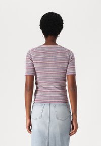 Une personne aux cheveux courts et bouclés porte une chemise à rayures à manches courtes et une jupe en denim léger, vue de dos sur un fond uni.