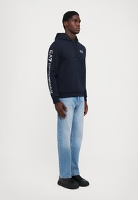 Felpa con cappuccio blu navy in cotone, dotata di tasca frontale e testo stampato bianco lungo la manica sinistra. Indossata con jeans di denim chiaro.