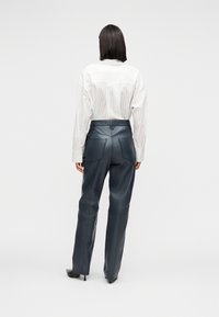 Femme aux cheveux bruns raides, portant une chemise blanche à rayures rentrée dans un pantalon en cuir noir taille haute et des talons noirs, debout devant un fond blanc.