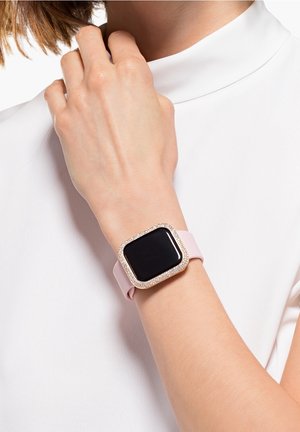 Personne portant une montre connectée avec un bracelet rose et un cadre doré orné de strass au poignet, main reposant près de la clavicule.