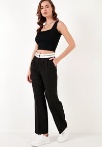 Črni skrajšani top v kombinaciji z črnimi hlačami, ki imajo bel pas. Model nosi bele superge; ozadje je enobarvno belo.