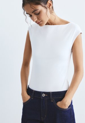 Femme aux cheveux foncés attachés, portant un haut blanc sans manches ajusté et un jean bleu foncé, les mains dans les poches, regardant vers le bas.