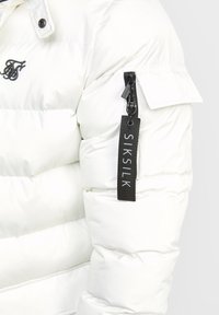 Vit vadderad jacka med horisontell quiltning, med en svart dragkedjeficka på vänster ärm och en märkt svart etikett. Vattentätt material.