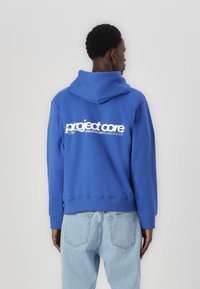 Sudadera con capucha azul con un gran texto gráfico en blanco "project core" impreso en la parte posterior. Tiene un ajuste holgado y puños acanalados.