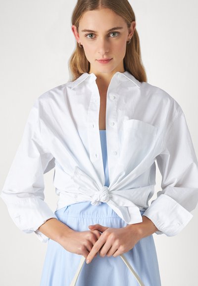 Cămașă albă cu nasturi, cu un nod în față, purtată peste o rochie de culoare albastru deschis. Are un singur buzunar �și mâneci lungi. Material din bumbac neted.