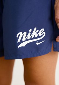 Mörkblå shorts med vit Nike-logotyp som bärs på en persons lår, med en hand vilande bredvid shortsen.