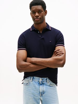 Man med korslagda armar i en marinblå polo med randig krage och ljusblå jeans som står mot en plain vit bakgrund.