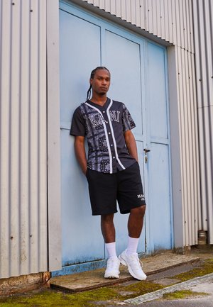 Short-sleeve black shirt with paisley pattern, white accents, and button front, paired with black shorts and white sneakers, standing by blue door.